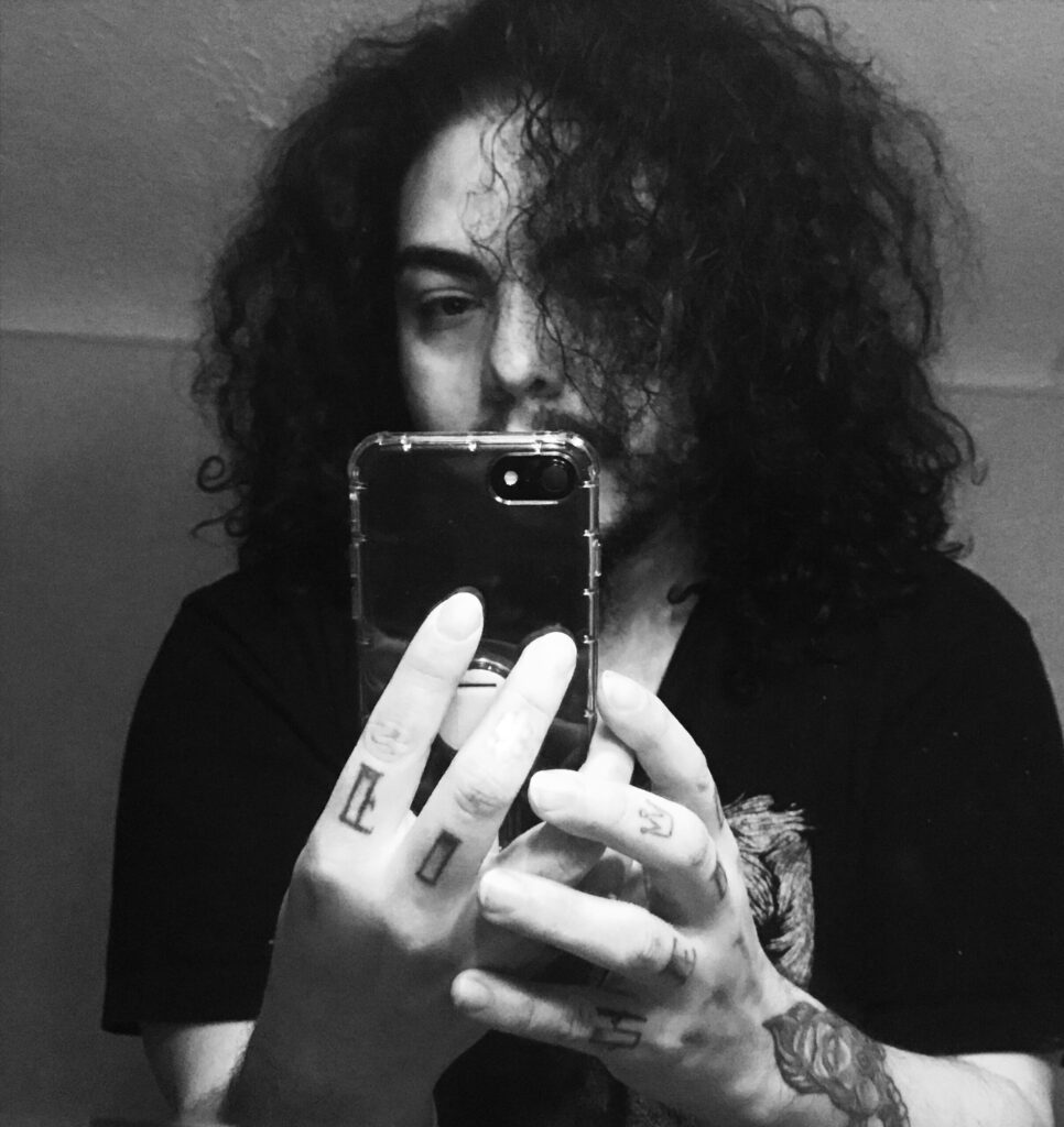 A Romani man with very curly long dark hair, dark eyebrows and facial hair - manicured - taking a selfie in a mirror. It's in black and white. His tattoos are visible on fingers and arm.
